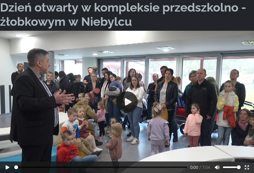 Zdjęcie przedstawia Dzień otwarty w kompleksie przedszkolno-żłobkowym w Niebylcu. Na obrazku widać, że odbywa się wydarzenie w przedszkolu. W pomieszczeniu jest kilka dorosłych i dzieci. Jeden z mężczyzn przemawia do publiczności. Wydaje się, że jest to nowoczesne pomieszczenie z białymi ścianami i oświetleniem. Na podłodze widać kolorowe zabawki lub materiały edukacyjne dla dzieci.