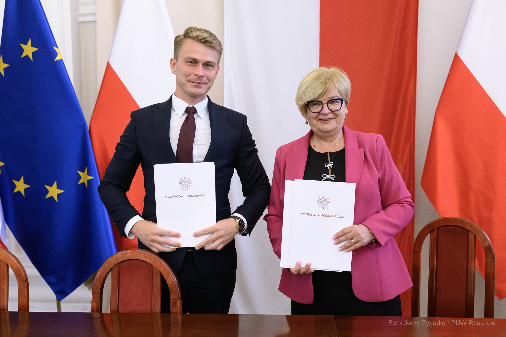 Dwoje osób stoi obok siebie, trzymając w dłoniach teczki z napisem ‘Wojewoda Podkarpacki’. W tle widoczne flagi Unii Europejskiej i Polski. Fot. Jerzy Żygadło / PUW Rzeszów