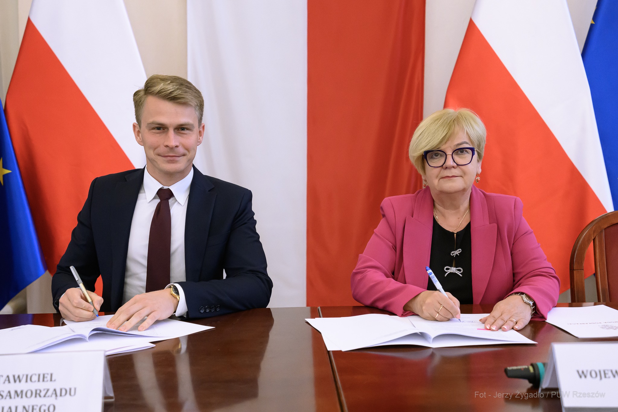 Dwoje osób siedzi przy stole i podpisuje dokumenty. Na stole otwarte teczki, w tle flagi Polski i Unii Europejskiej. Fot. Jerzy Żygadło / PUW Rzeszów.